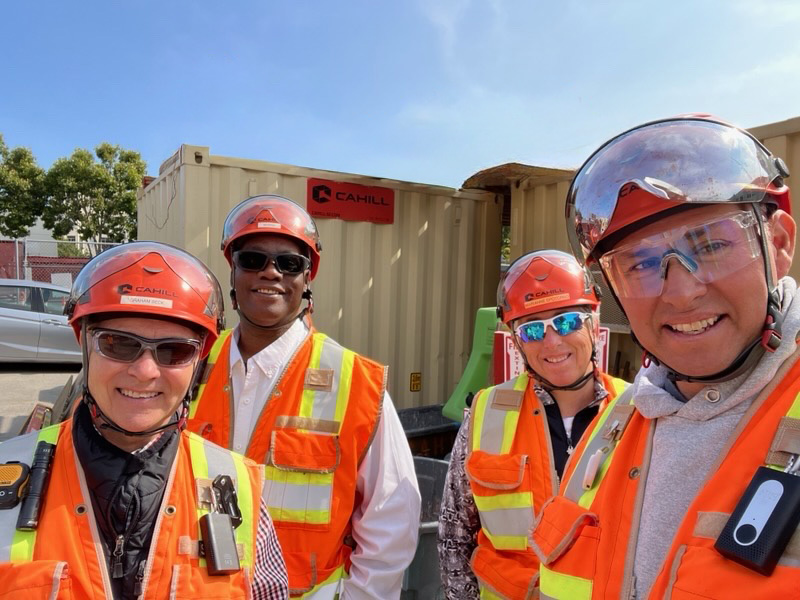 Four construction workers wearing orange safety vests and hard hats, smiling outdoors near a shipping container labeled Cahill.