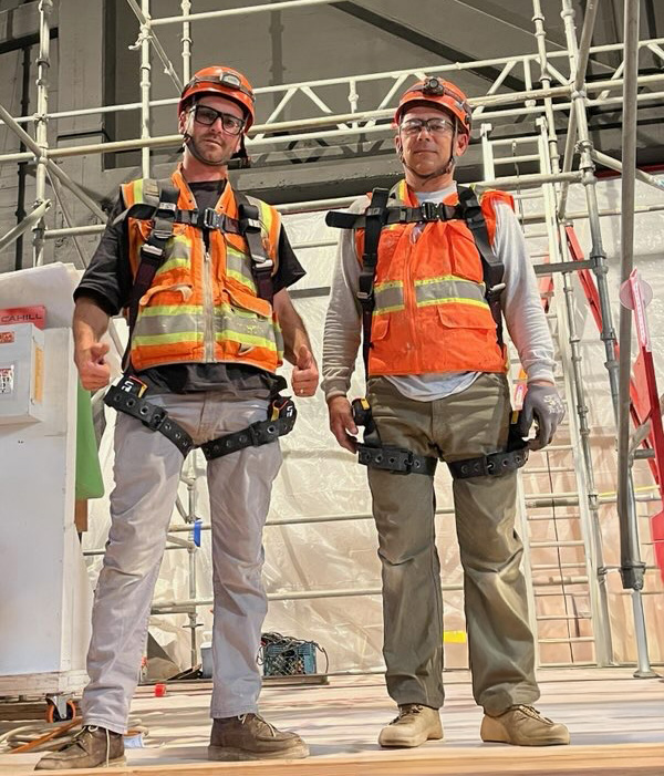 Two construction workers wearing orange safety vests, helmets, harnesses, and work boots standing inside a construction site with scaffolding.