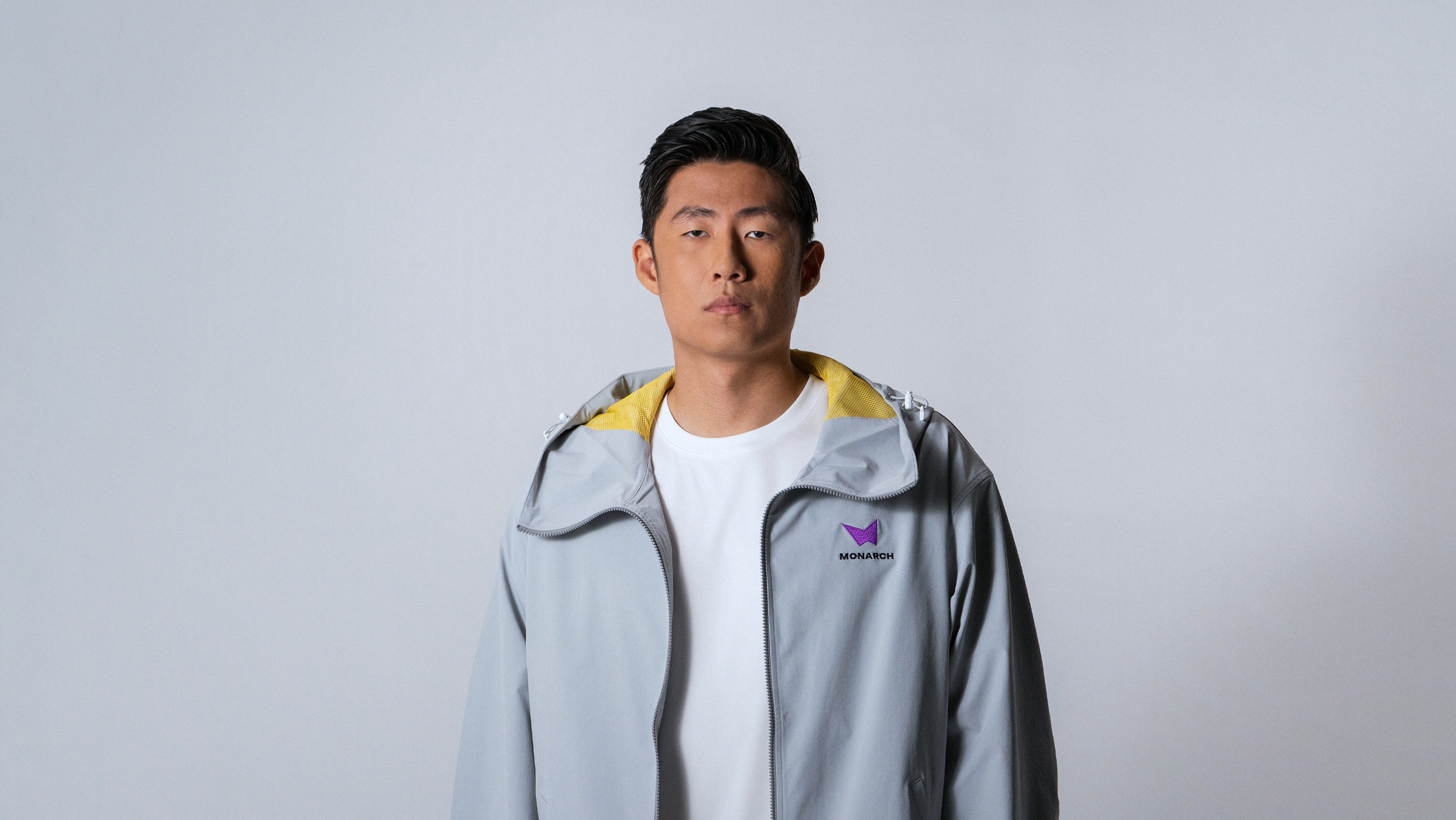 Man wearing a light gray jacket with a yellow inner lining and the Monarch logo over a white shirt, standing against a plain gray background.