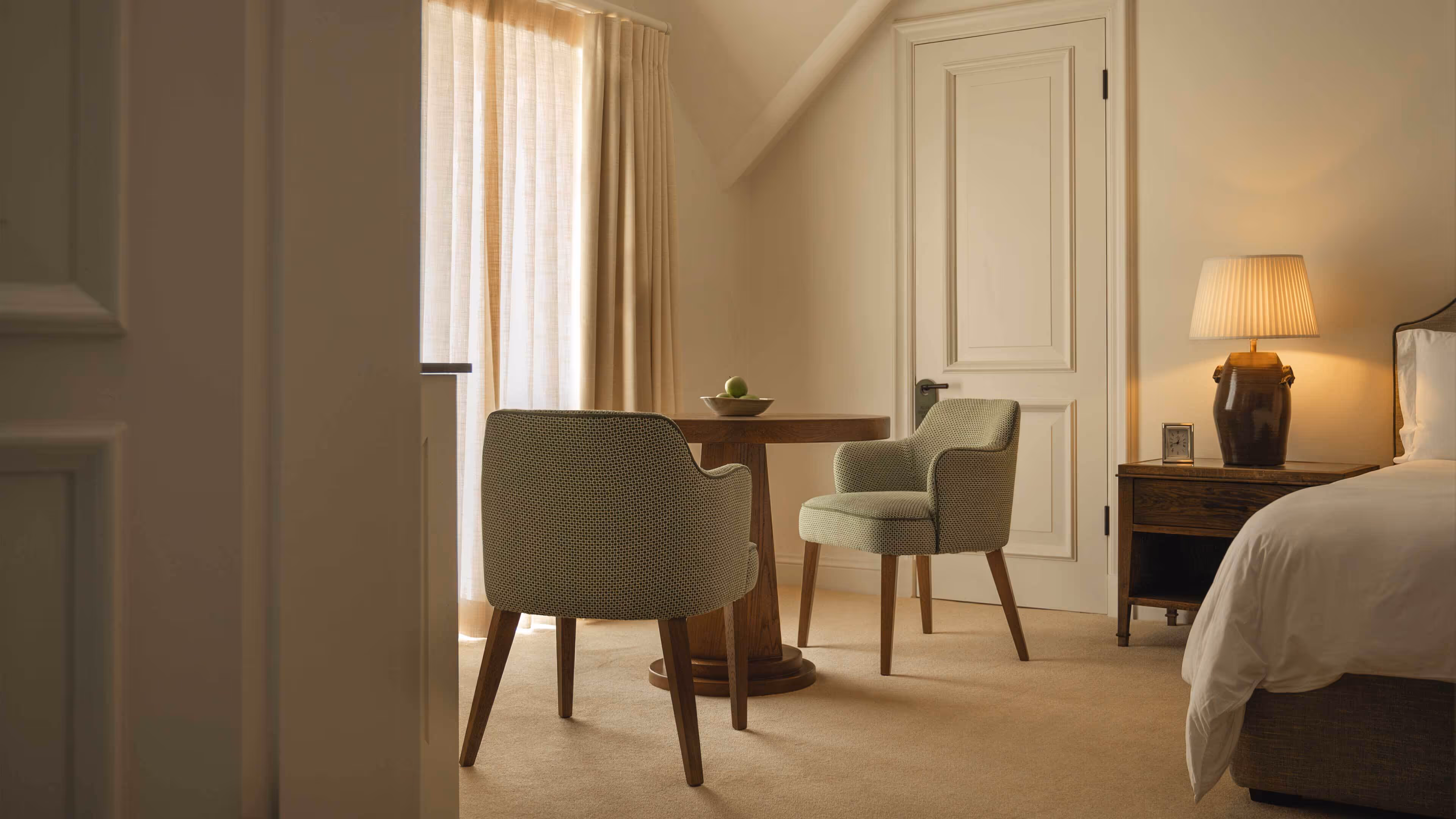 Cozy bedroom corner with two upholstered chairs around a wooden round table, next to a nightstand with a lamp and a door in the background.