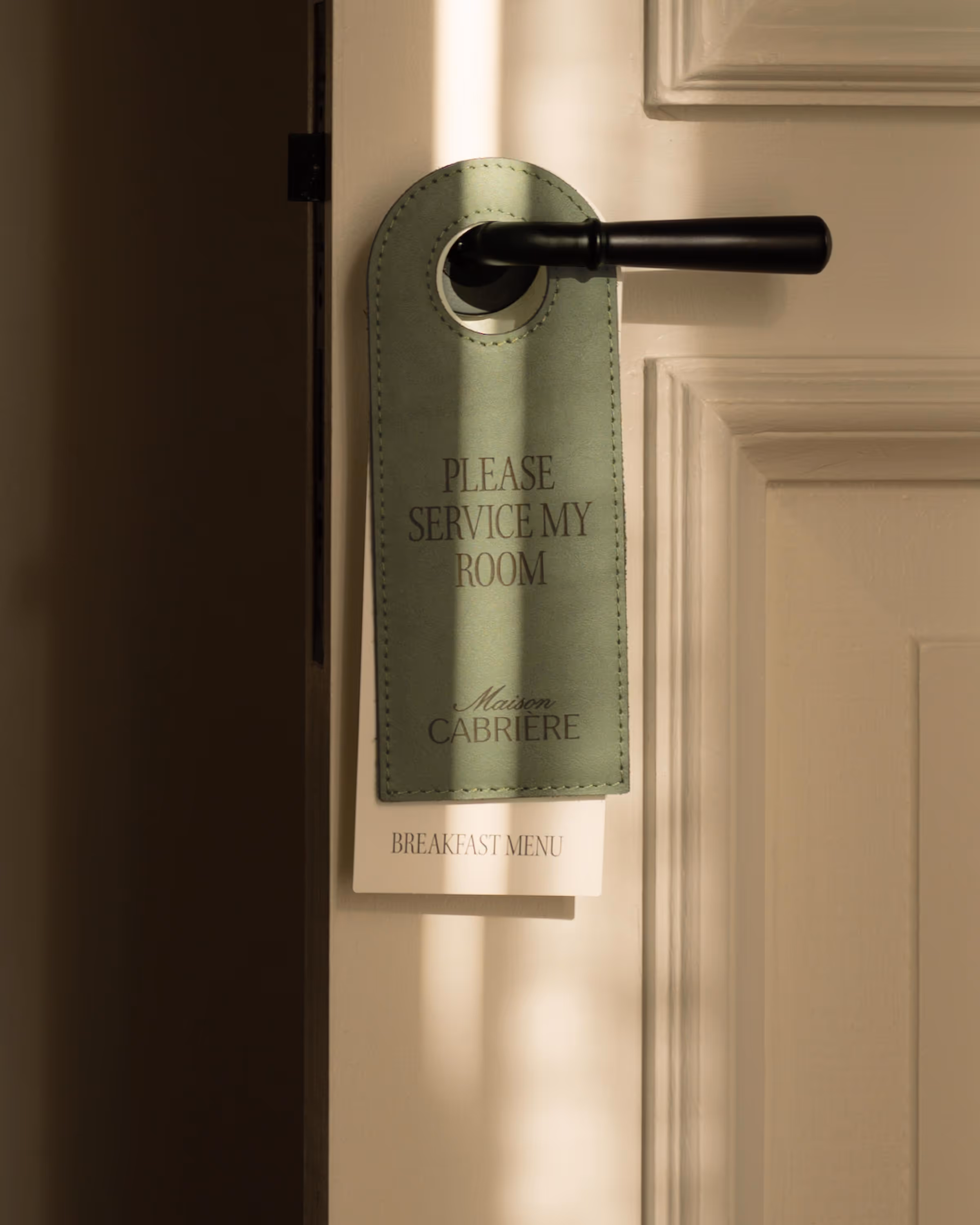 Green door hanger with text 'Please Service My Room' and 'Maison Cabrière' hanging on a white door handle with a breakfast menu underneath.