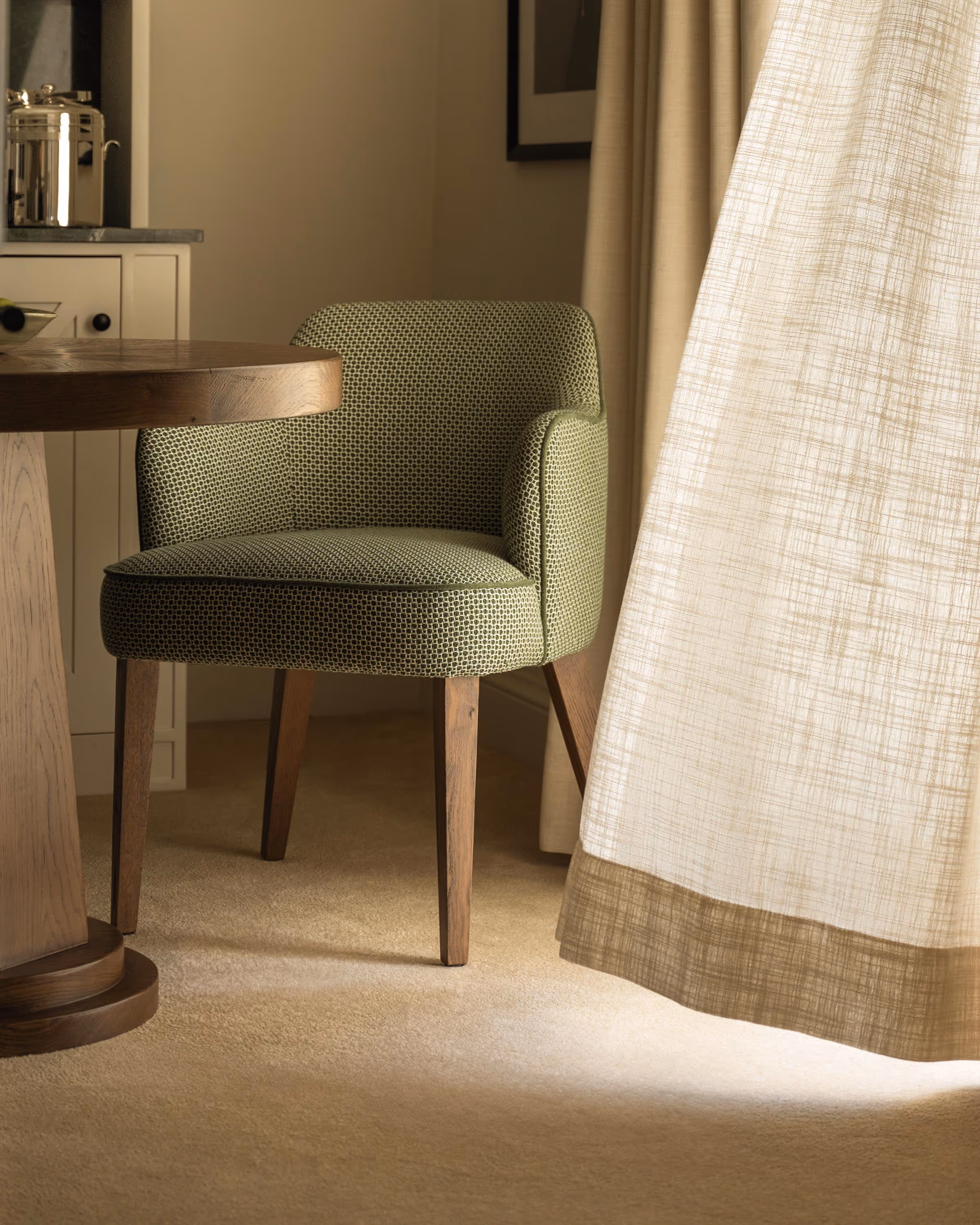 Green upholstered chair with wooden legs next to a wooden table and beige curtains illuminated by soft light.