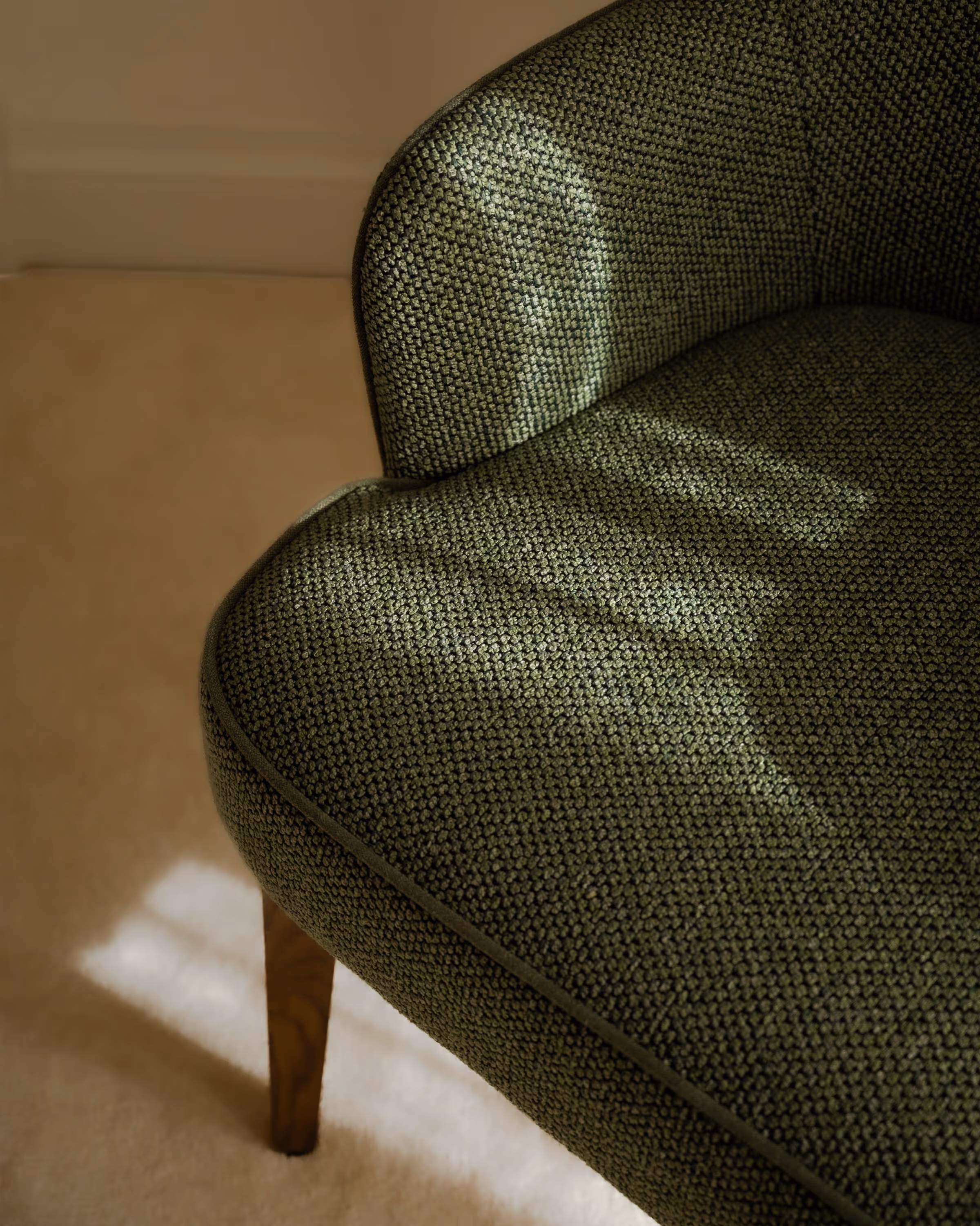 Close-up of a green textured upholstered chair leg on a beige carpet with sunlight casting shadows.