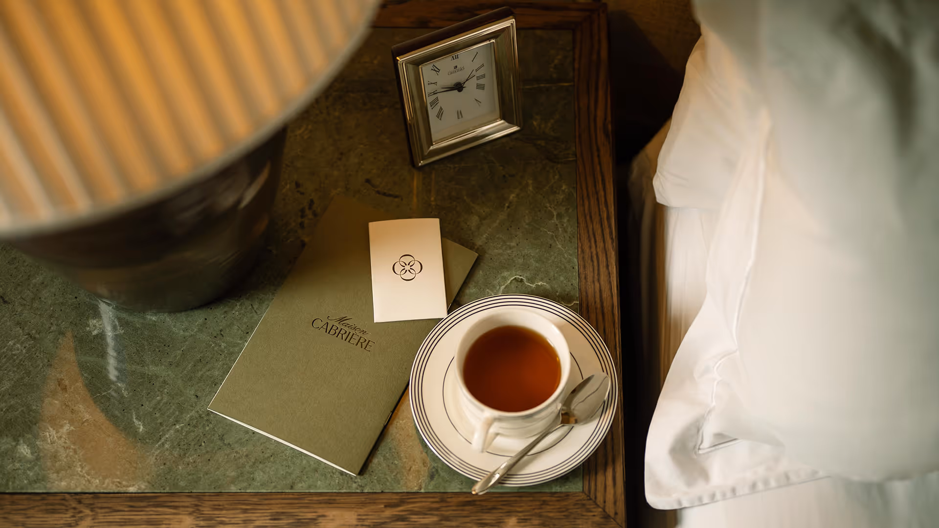 A bedside table with a cup of tea, a spoon on a saucer, a green booklet labeled 'Maison Cabriere,' a small card, a silver-framed clock, and a lamp base next to a white pillow.