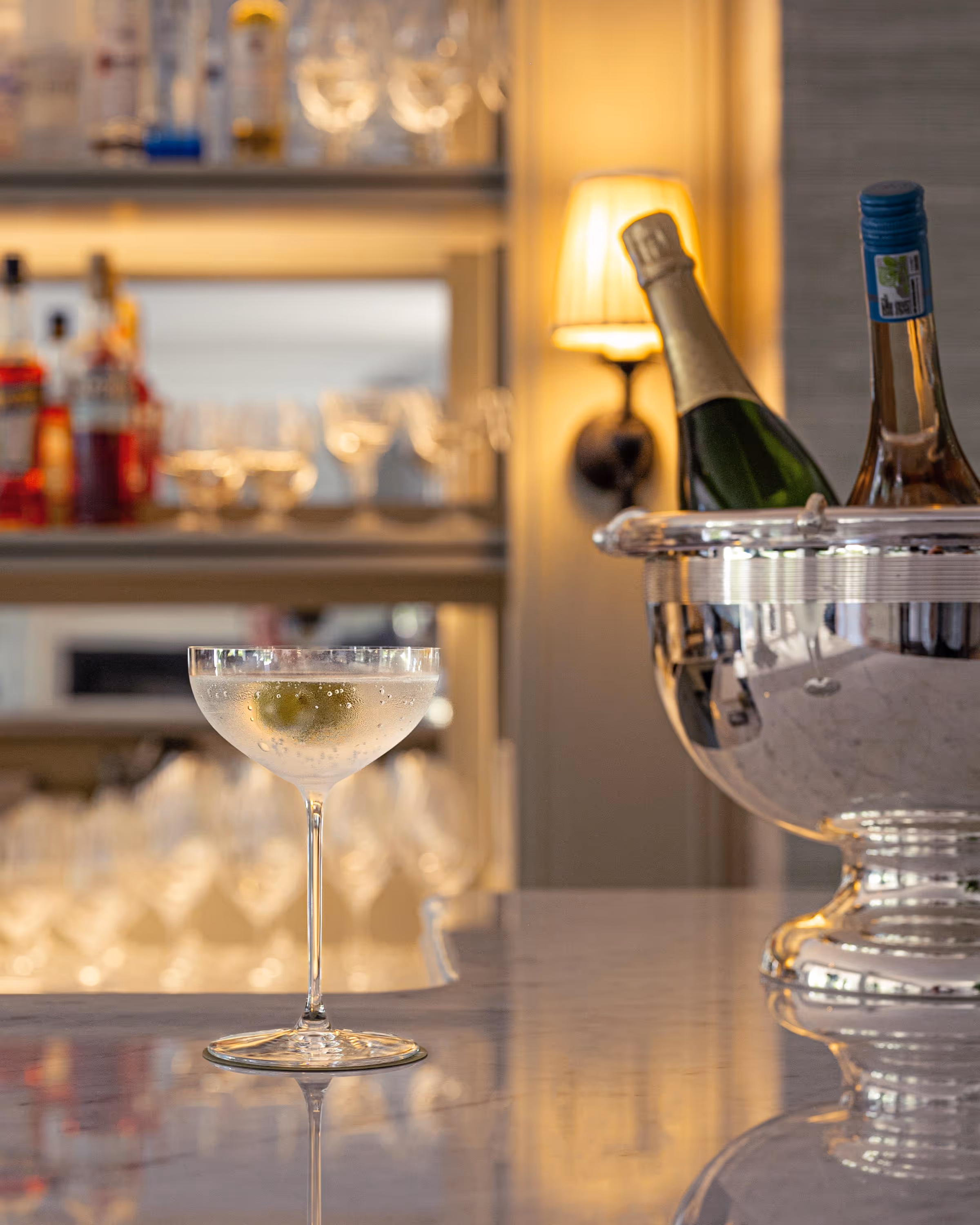 Chilled cocktail glass with an olive on a bar counter next to a silver ice bucket holding a champagne and white wine bottle.