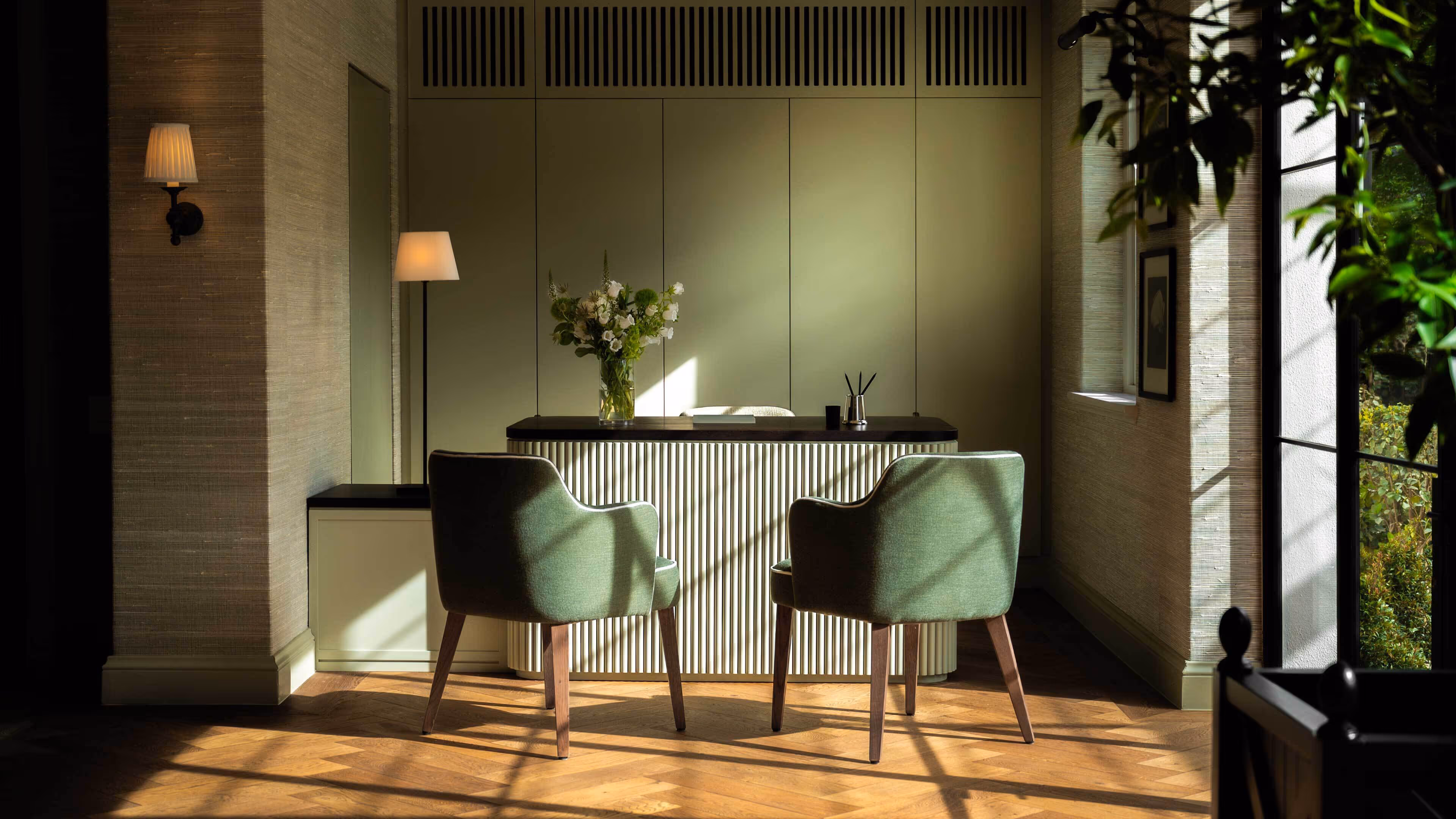 Two green upholstered chairs in front of a modern desk with a flower vase in a softly lit room with wooden flooring and large windows.