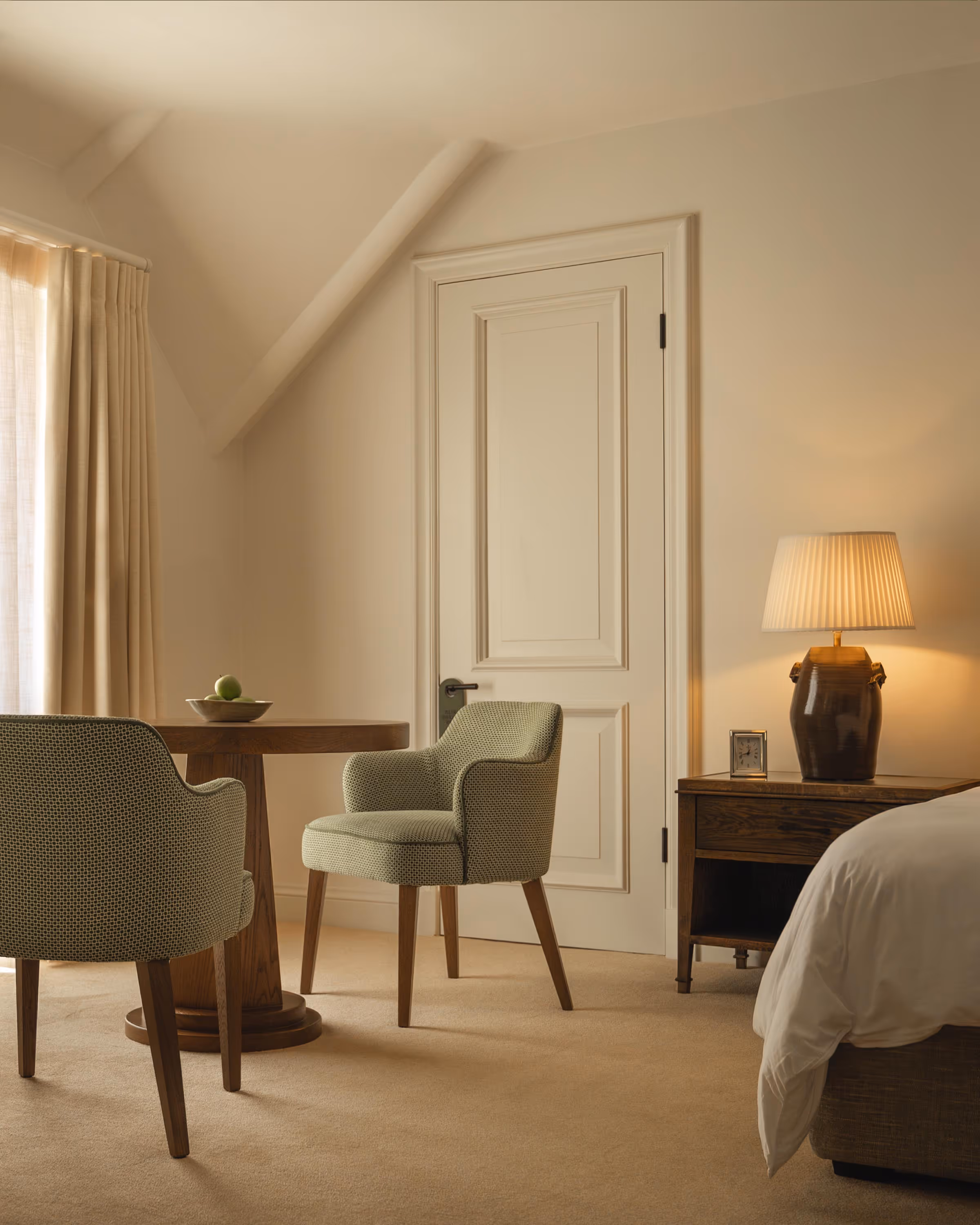 Cozy bedroom corner with beige carpet, two green upholstered chairs around a wooden round table with a bowl of green apples, a nightstand with a lit lamp and a small clock, and a closed white door.