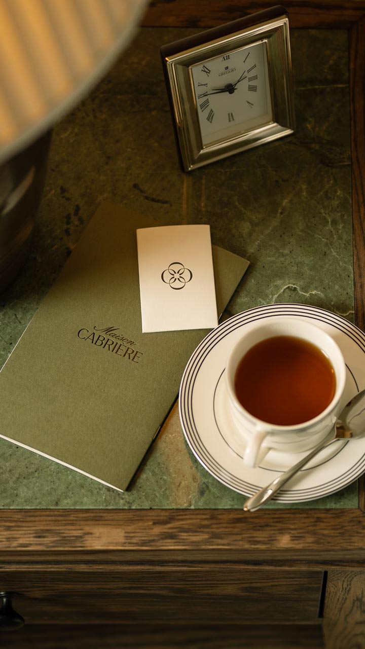A cup of tea on a white saucer with silver stripes next to a teaspoon, a green Maison Cabriere booklet, a small card with a flower-like logo, and a silver-framed clock showing 8:00 on a marble tabletop.