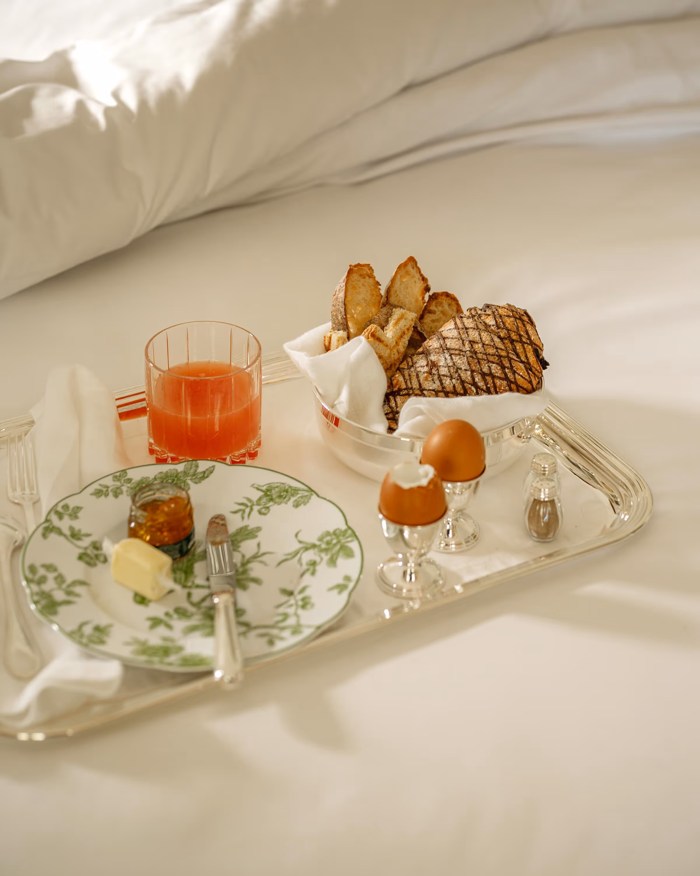 Breakfast tray on a bed with two soft-boiled eggs in silver holders, a bowl of sliced bread, a glass of orange juice, a plate with butter and jam, a knife, and a fork.