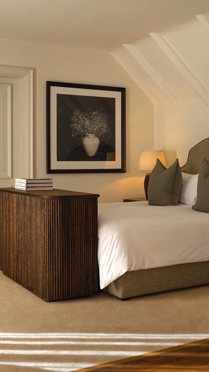 Cozy bedroom with a beige bed, green pillows, wooden furniture, a lamp, and framed artwork of a vase with flowers.