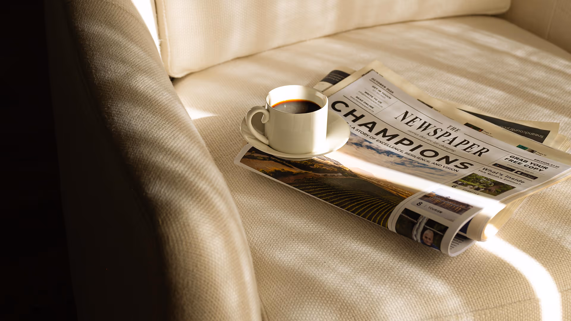 White cup of black coffee on a saucer placed on top of a folded newspaper on a beige fabric chair.