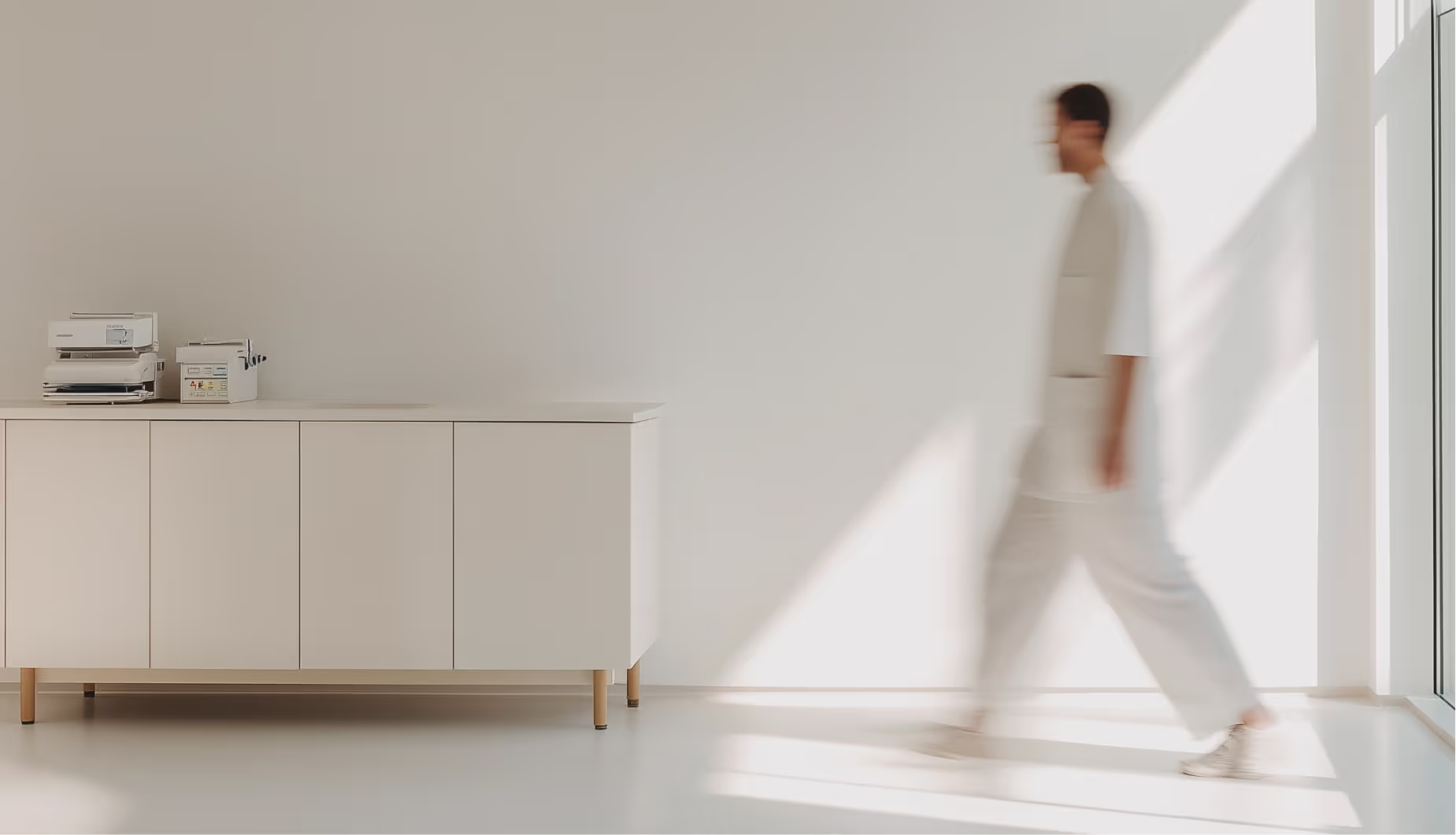 Rozmazaná osoba v bílém oděvu kráčí v minimalistické světlé místnosti vedle bílého nábytku s kancelářskými přístroji.