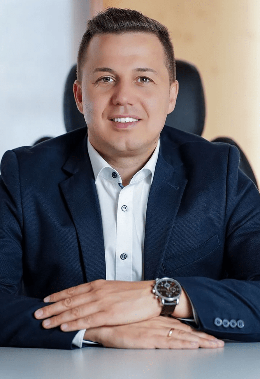 Ein Mann im dunklen Sakko mit weiĂem Hemd sitzt an einem Tisch, lĂ€chelt in die Kamera und hat die HĂ€nde entspannt vor sich verschrĂ€nkt. Im Hintergrund ist ein unscharfer BĂŒrostuhl sichtbar.