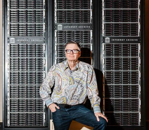 Mark Graham, wearing glasses and a paisley shirt, stands in front of three large, blinking internet servers in Internet Archive.