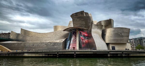 A long view of the billowing forms of the museum, sitting on the edge of the water with storm clouds overhead.