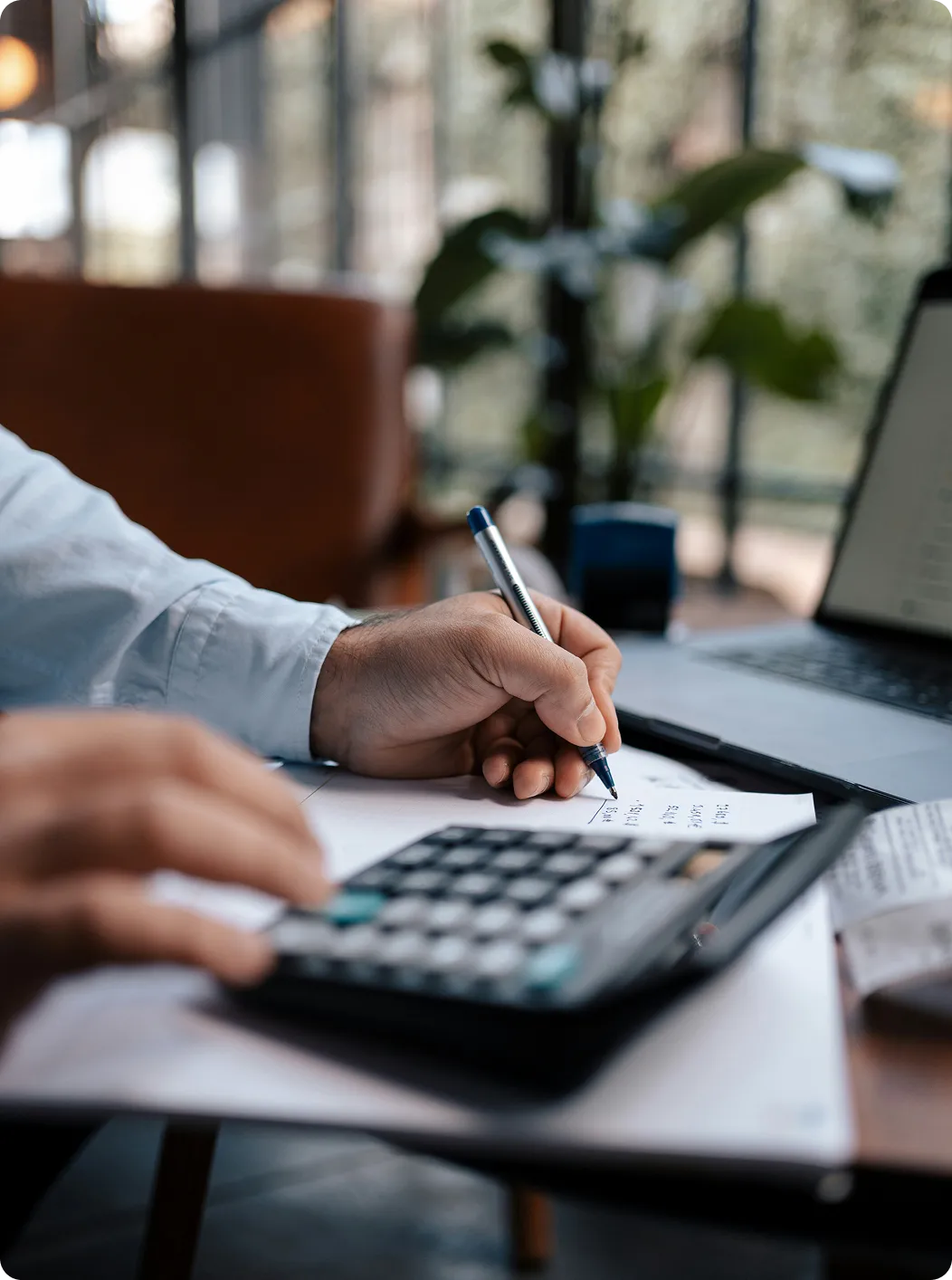 A man writing on a calculator with a laptop in the background.