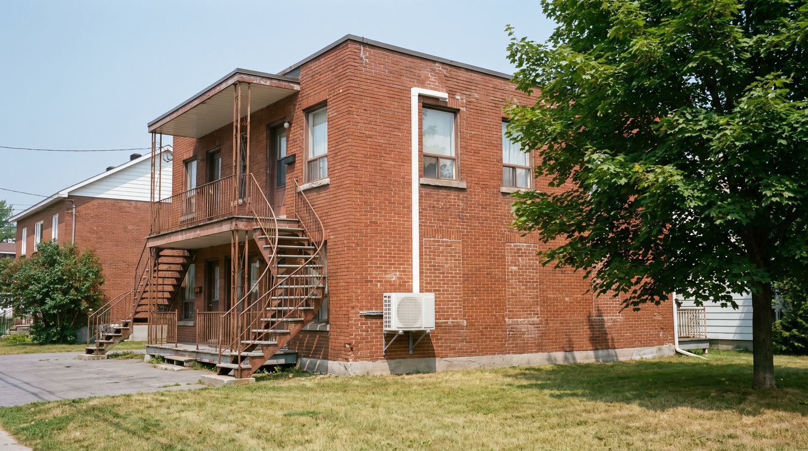 Unité extérieure de climatiseur mural fixée sous la fenêtre d'un duplex en brique de Gatineau par une journée chaude d'été