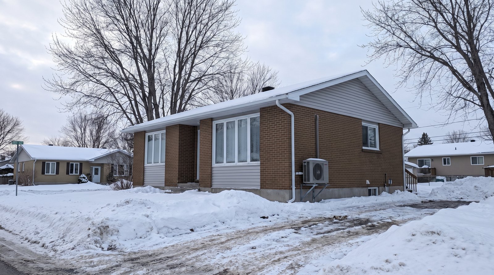 Bungalow en brique des années 1970 à Gatineau en hiver avec thermopompe murale sur socle surélevé au-dessus de la neige