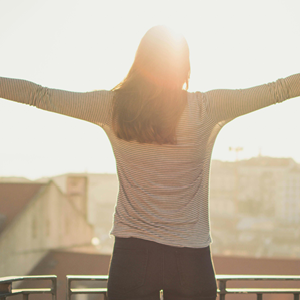 Person with long hair standing on a balcony with arms outstretched facing the bright sun.