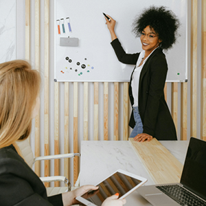 Two women in an office, one standing and writing on a whiteboard while smiling, the other seated holding a tablet.