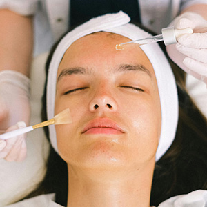 Woman with closed eyes receiving facial treatment with serum dropper and brush application.