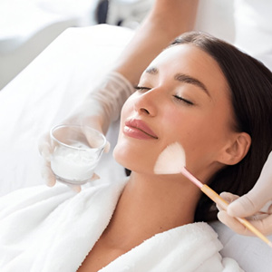Woman with eyes closed receiving a chemical peel treatment on her face from a professional wearing gloves.