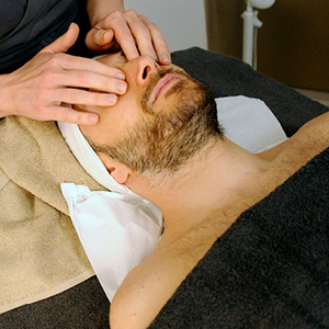 Man with beard receiving a facial massage while lying with a towel under his head and a black blanket covering his body.