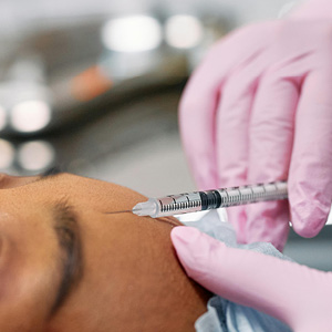 Close-up of a gloved hand holding a syringe injecting the forehead of a person lying down, representing a cosmetic neuromodulator treatment.