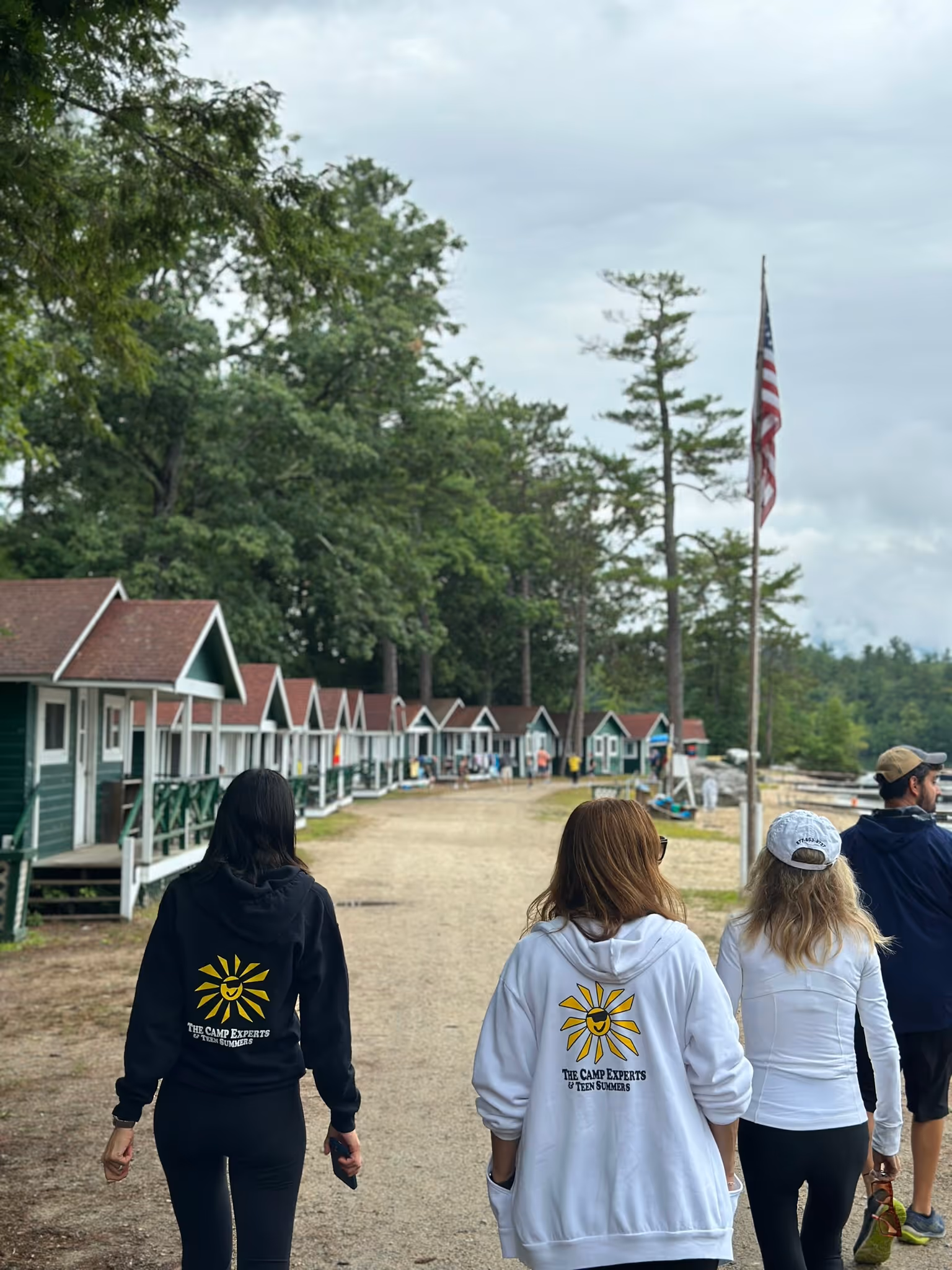 Four people walking away on a dirt path lined with small green cabins, with one person wearing a white hoodie and another wearing a black hoodie featuring a yellow sun logo and the text 'The Camp Experts & Teen Summers'.