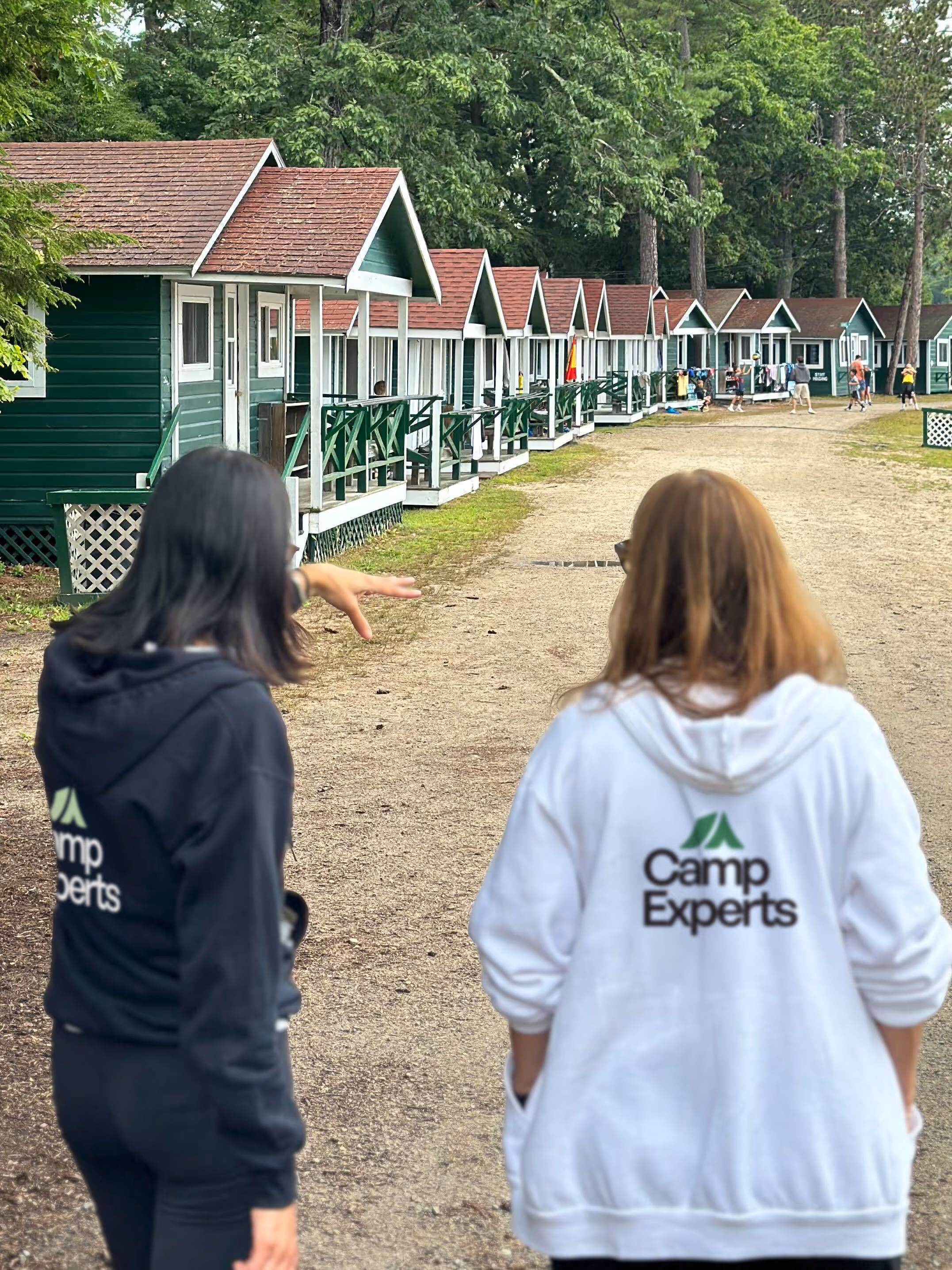 Two people wearing Camp Experts hoodies walking on a gravel path past a row of green cabins with red roofs surrounded by trees.