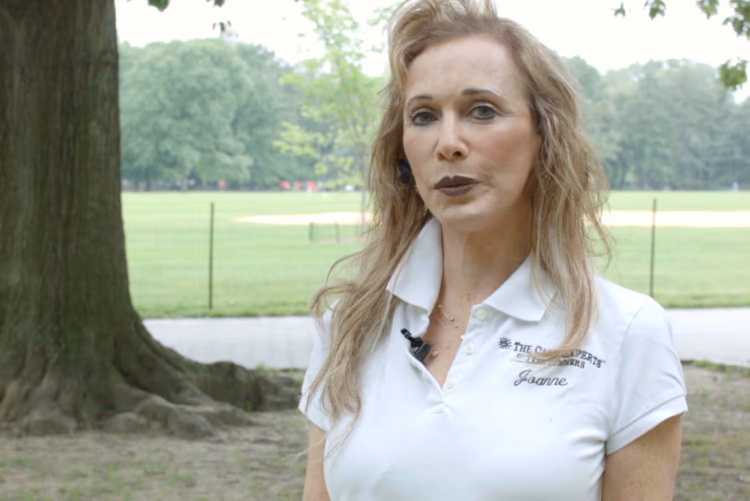 Woman with long blonde hair wearing a white polo shirt standing outdoors near a tree and a fenced grassy field.