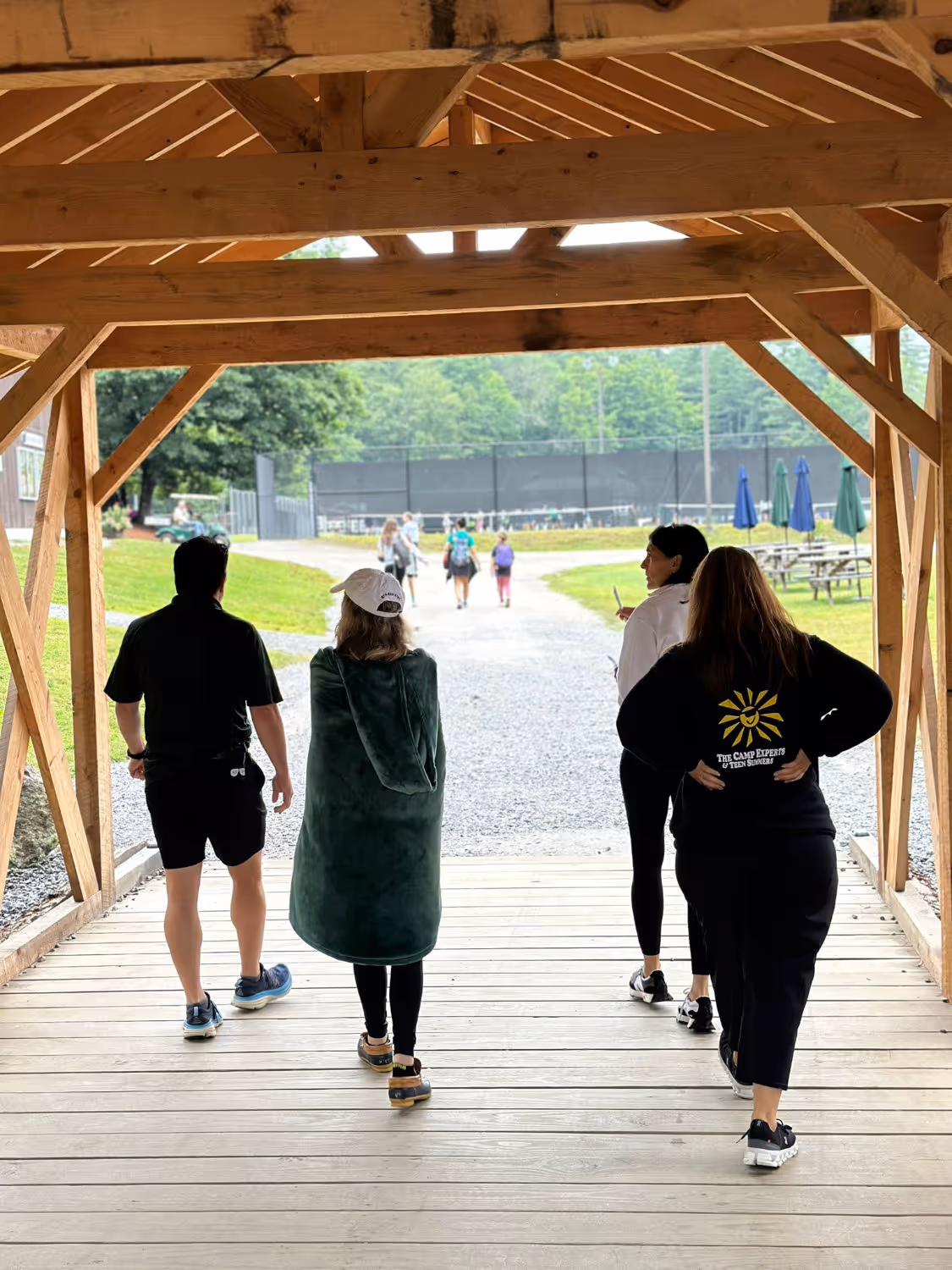 Four people walking under a wooden pavilion toward a gravel path with green trees and picnic tables in the background.