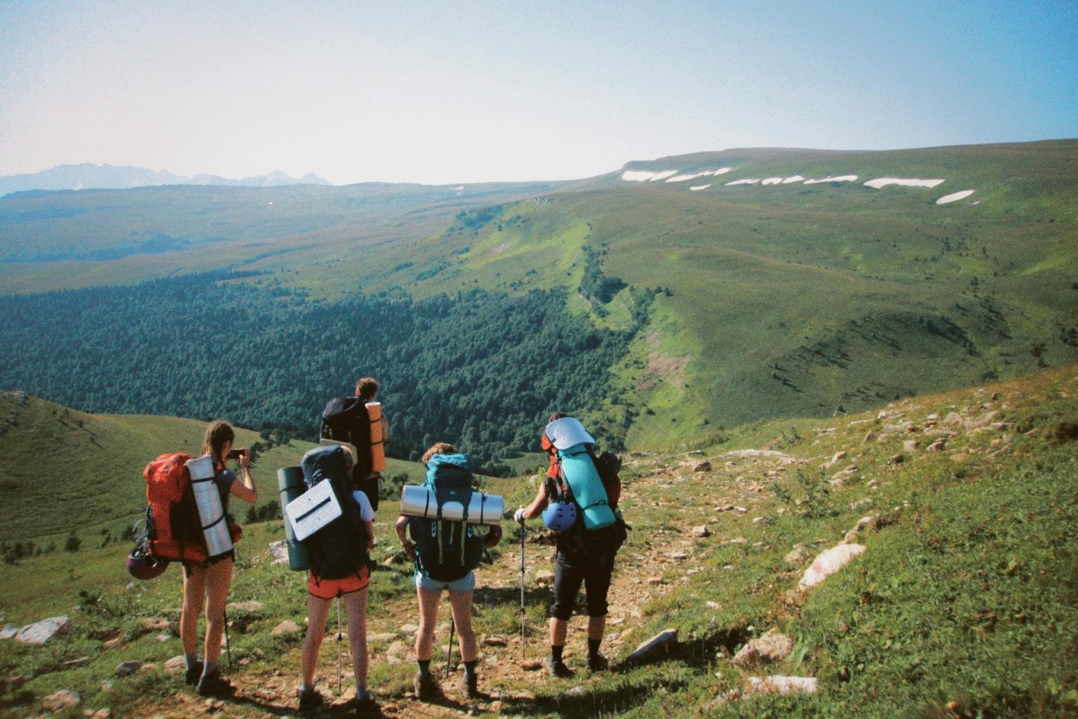 Group of four hikers with large backpacks standing on a rocky hillside overlooking a green valley and mountain range.