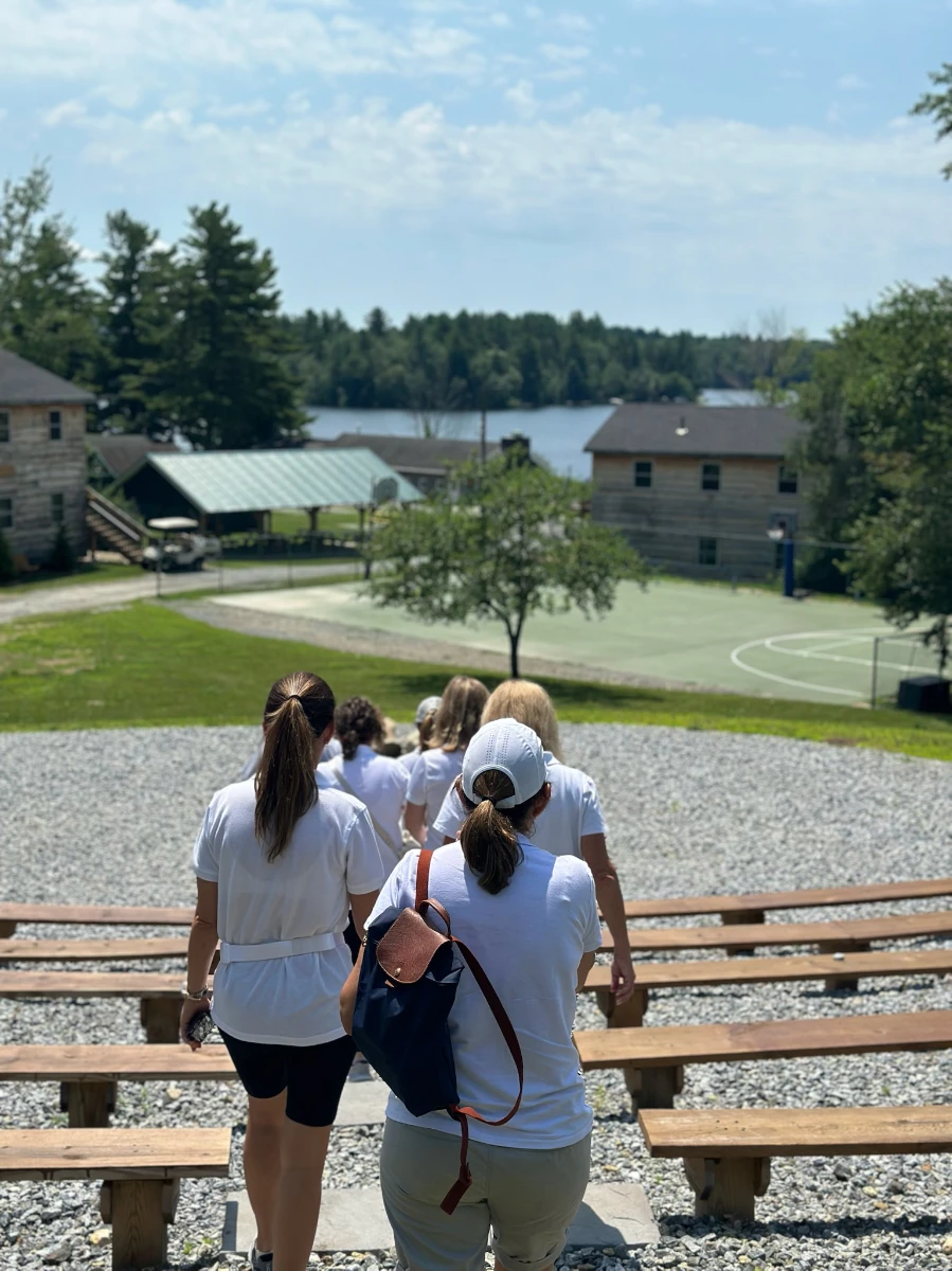 Group of people walking down wooden steps outdoors toward a lake and buildings with trees in the background.