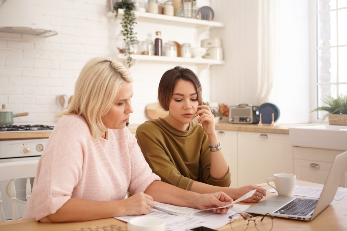 Two women sitting at a kitchen table reviewing documents with a laptop and coffee cups.
