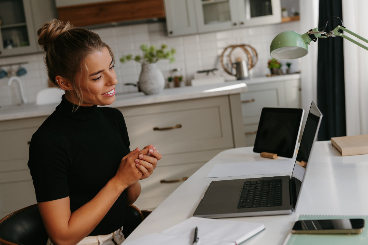 Young woman in black turtleneck sitting at a desk in a kitchen, smiling and engaging with an open laptop.