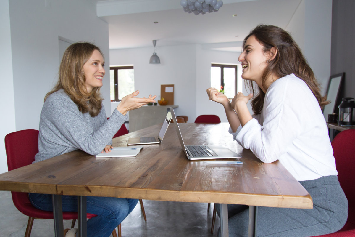 Two women sitting opposite each other at a wooden table, smiling and discussing with laptops open in front of them.