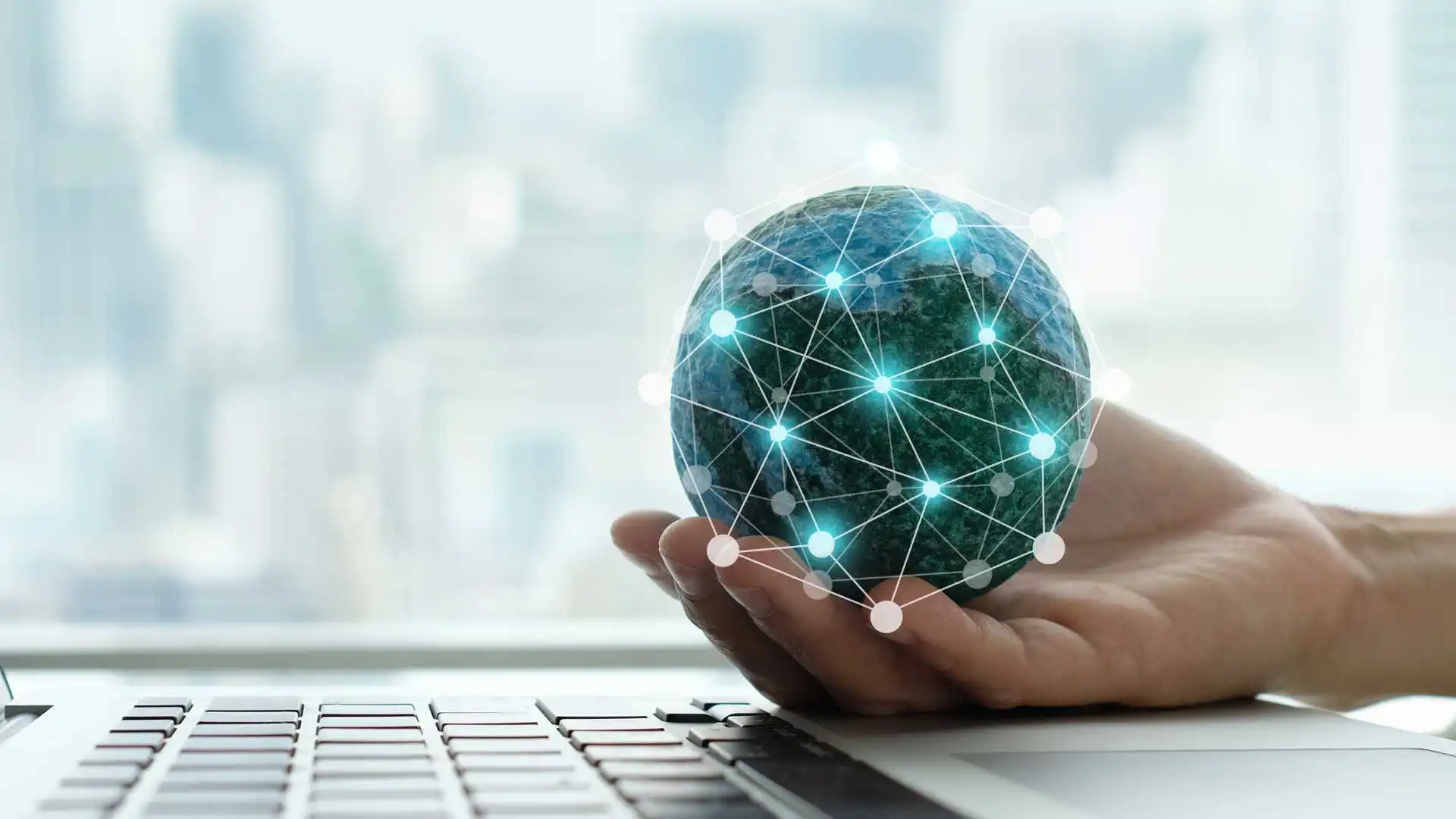 A hand holds the earth as a blue ball with green network in front of a computer