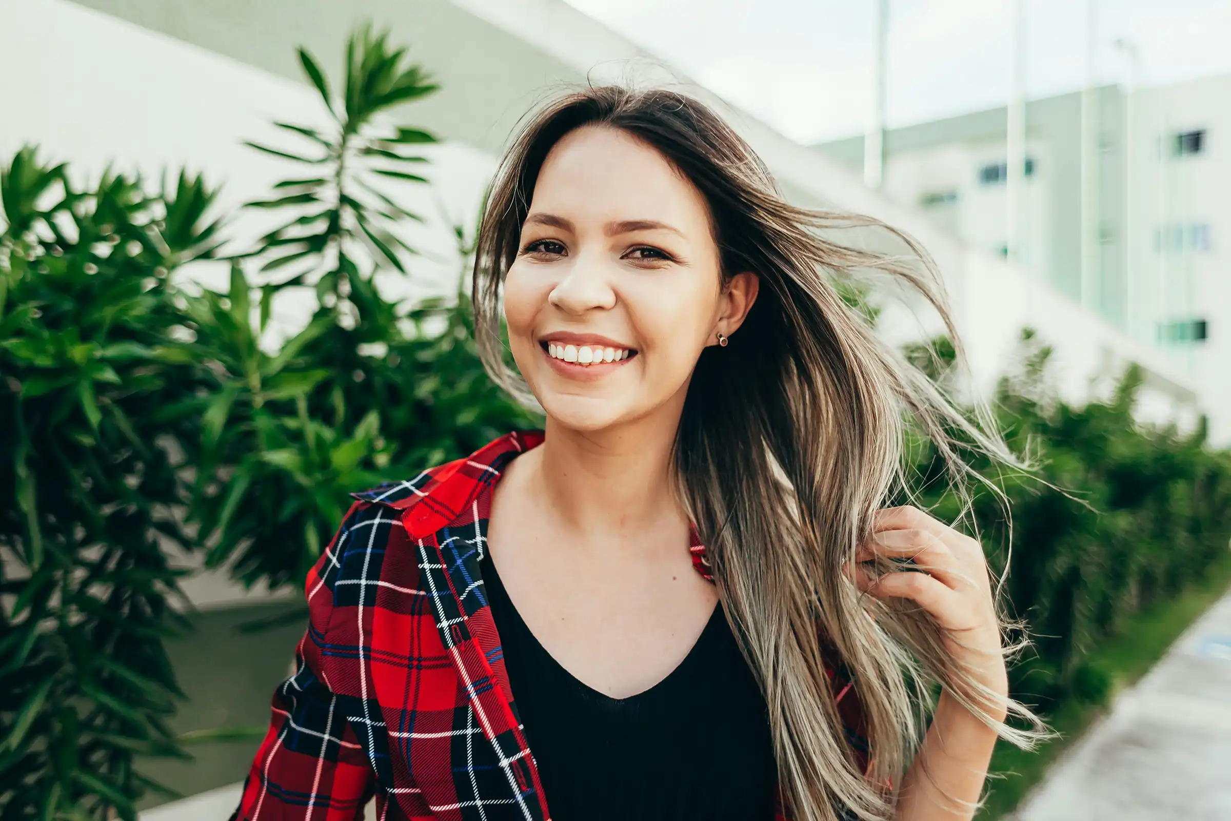 Smiling woman with long hair flowing in the breeze, wearing a red plaid shirt and standing outdoors, radiating clear skin, confidence, and natural energy.