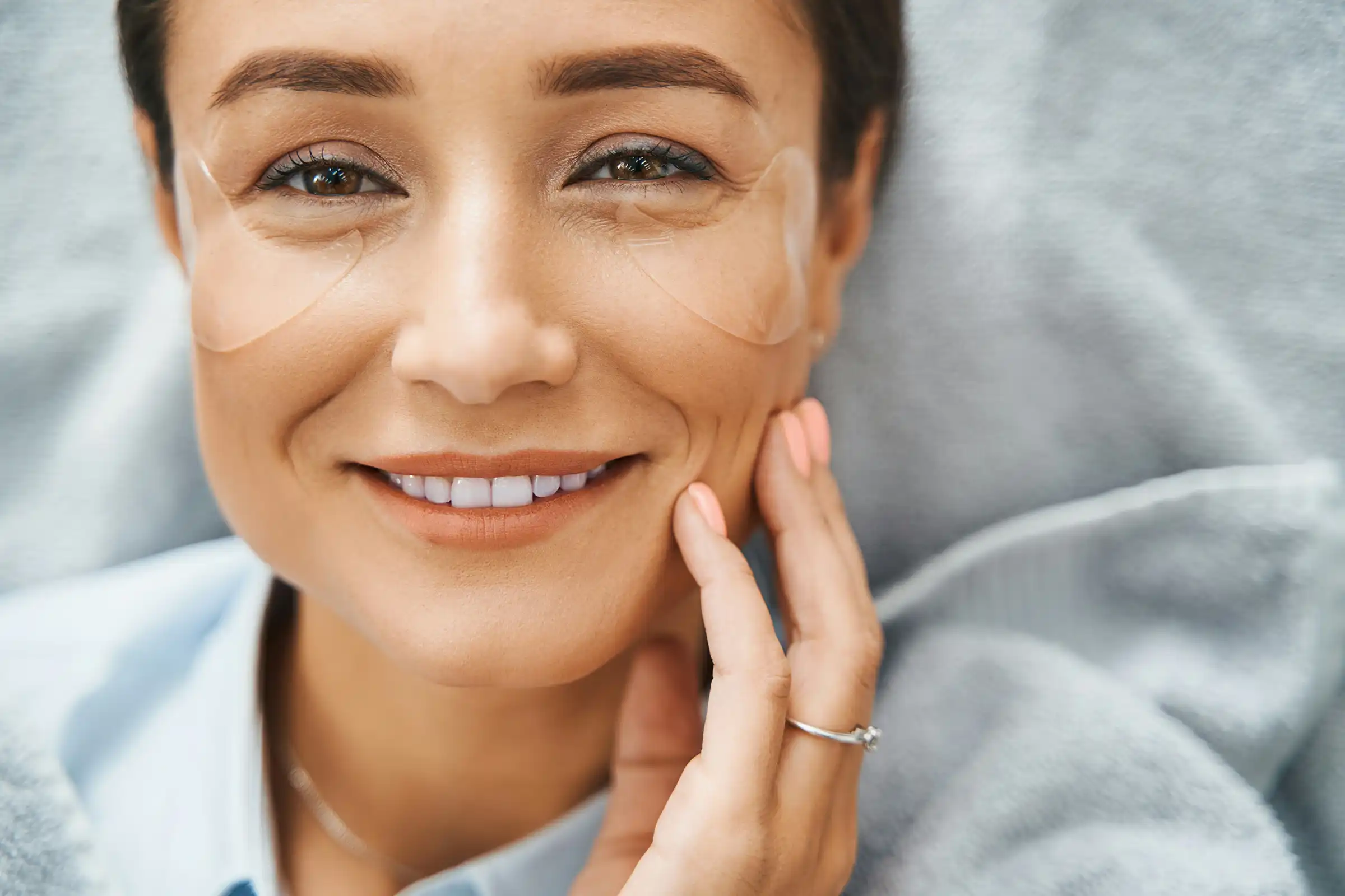 Woman smiling with bright eyes and hydrogel patches under her eyes, highlighting under-eye rejuvenation, smooth skin, and post-treatment radiance.
