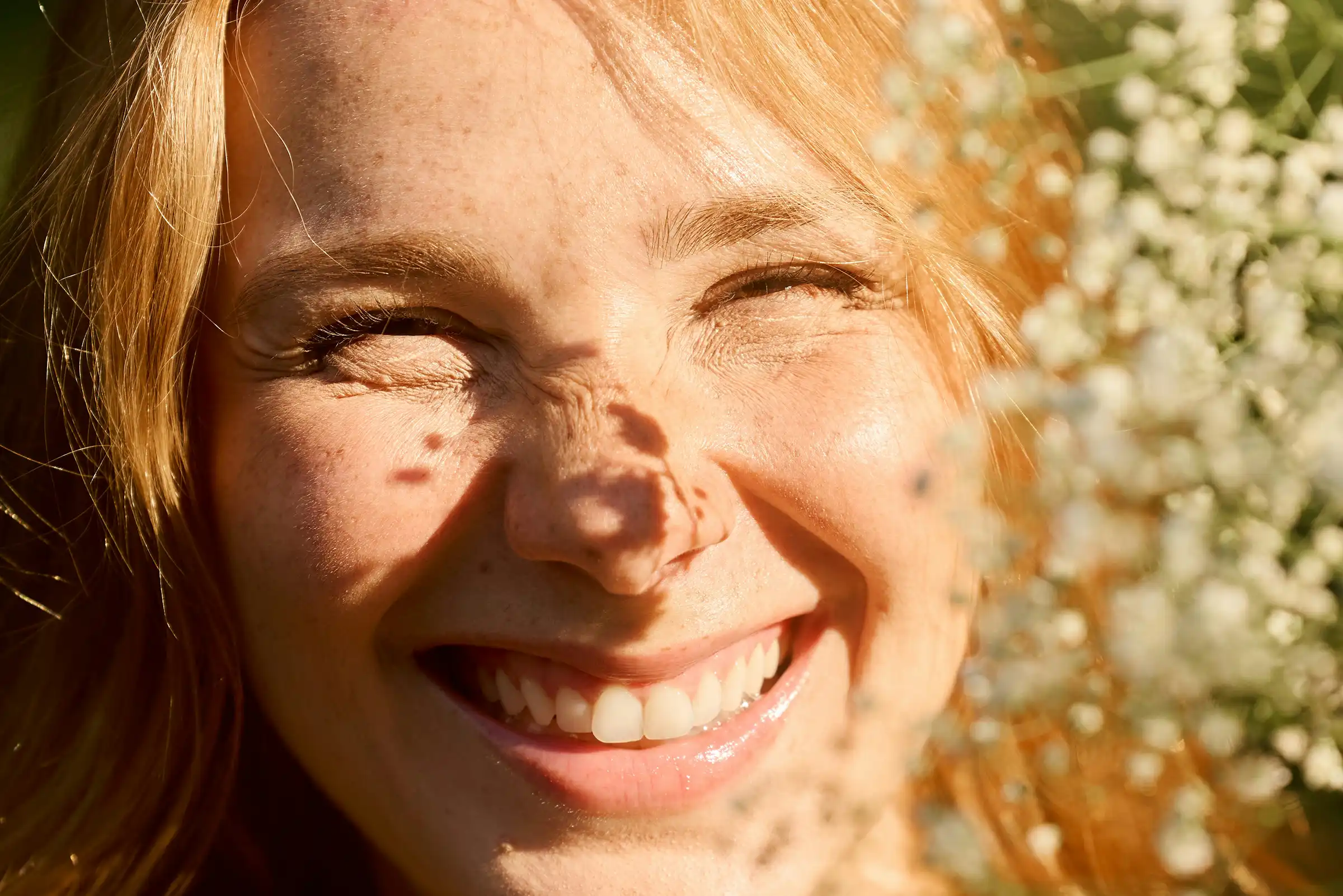 Close-up of a smiling woman with freckles beaming in the sunlight, partially framed by delicate white flowers—capturing radiant skin, joy, and natural beauty.
