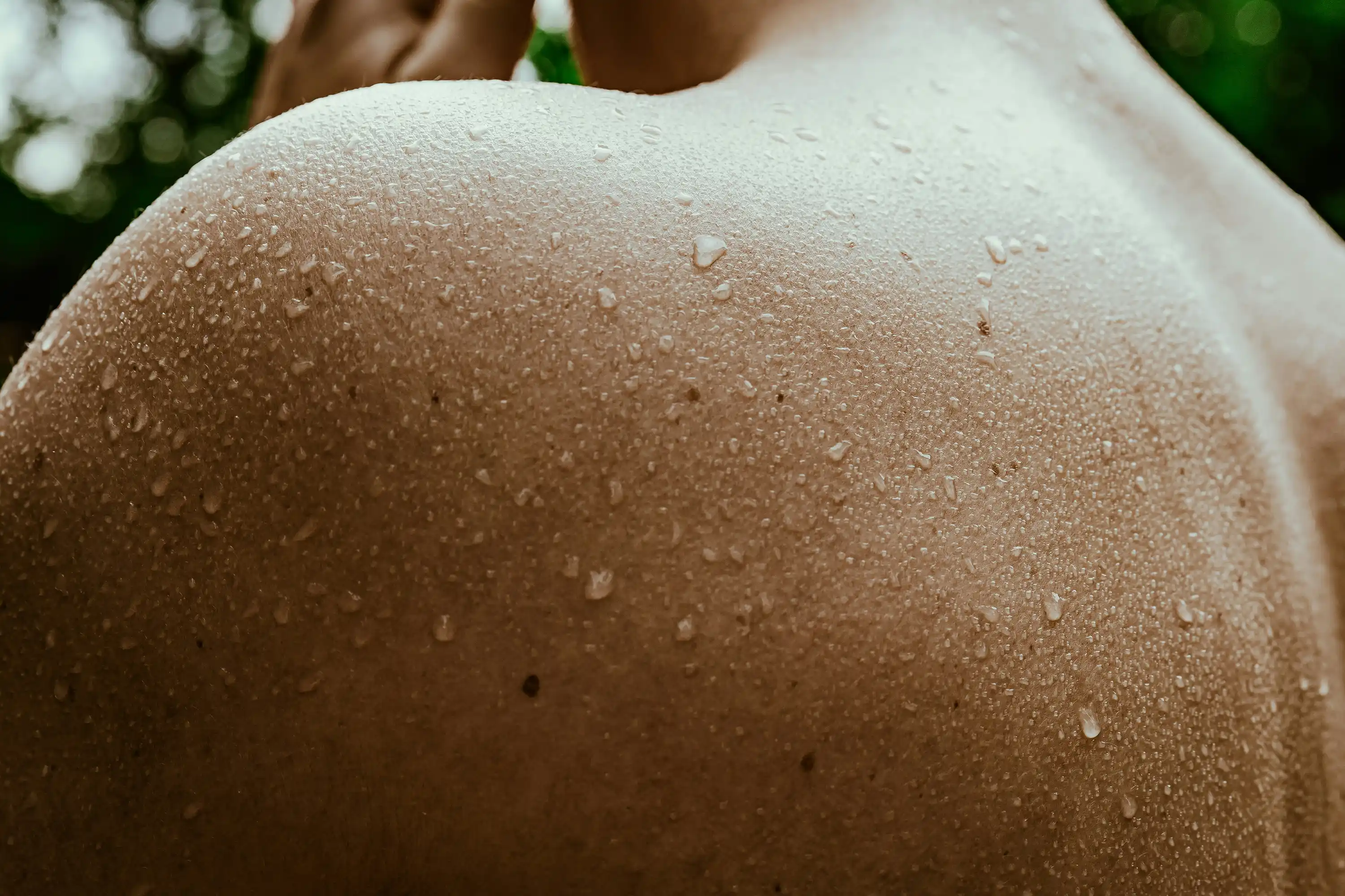 Close-up of a person’s bare upper back covered in fine water droplets, highlighting smooth, hydrated skin and natural texture in a serene, outdoor setting.
