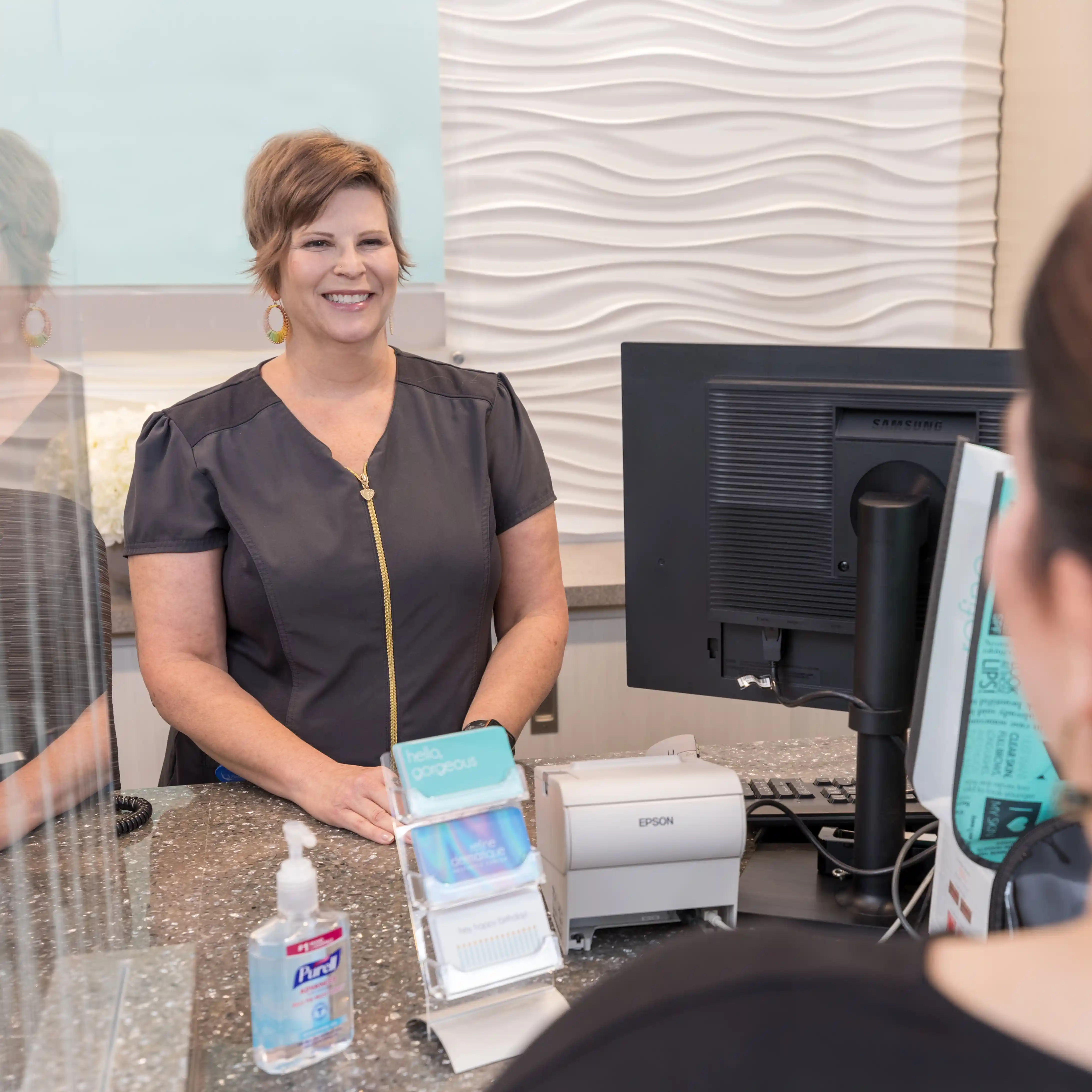 Friendly front desk staff member smiling at the reception area, representing warm patient welcome and exceptional service at Element Dermatology.