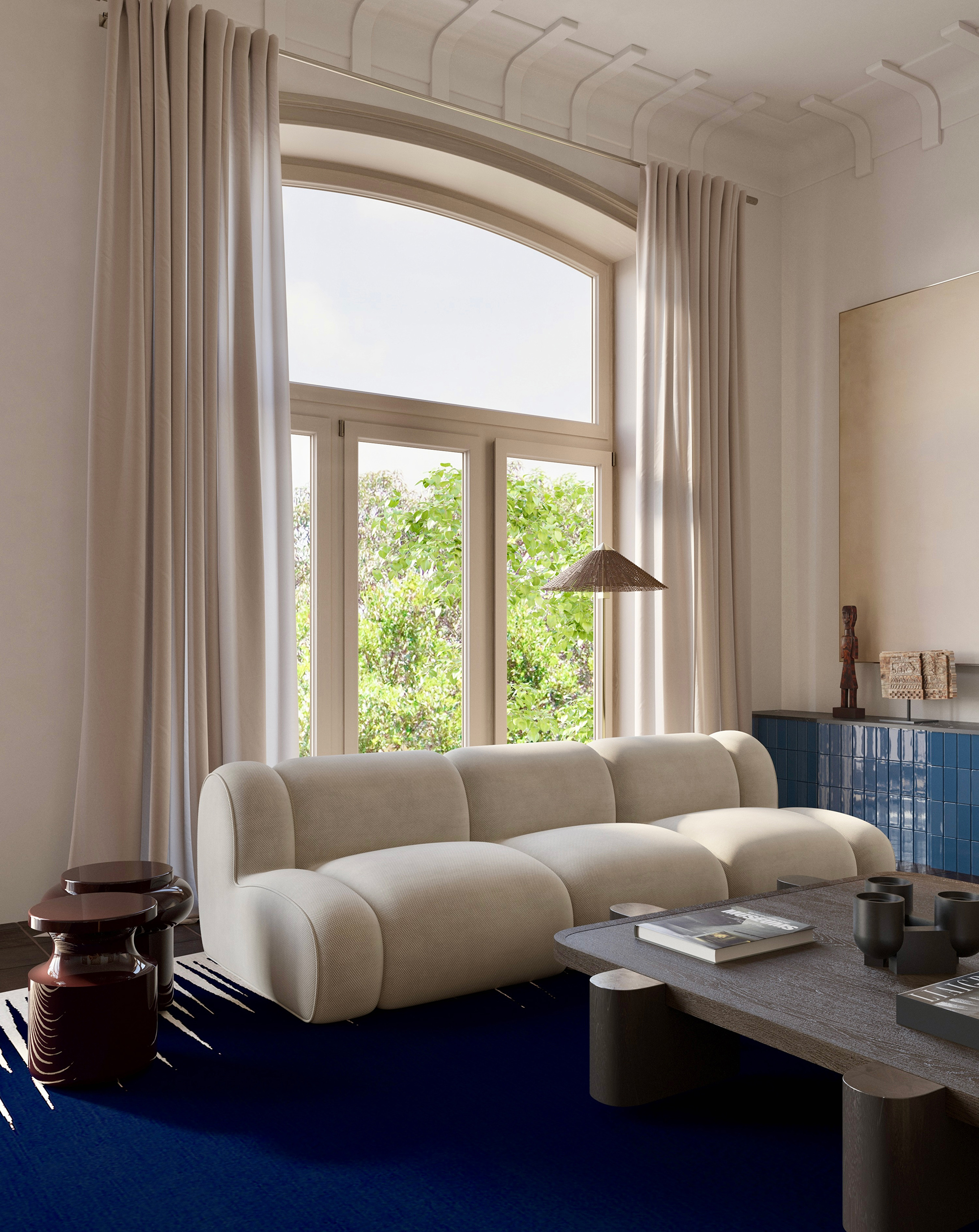 Bright living room with a sculptural white sofa Popus Edition Invisible collection  and a dark wood and ceramic custom coffee table. Large arched window with creamy drapes frames a view of lush greenery. Deep blue rug by Atelier Fevrier and glossy burgundy side tables by Delcourt Collection anchor the space with contrast and richness. Vintage Paavo Tynnel floor lamp from 1stdibs. {vertical}