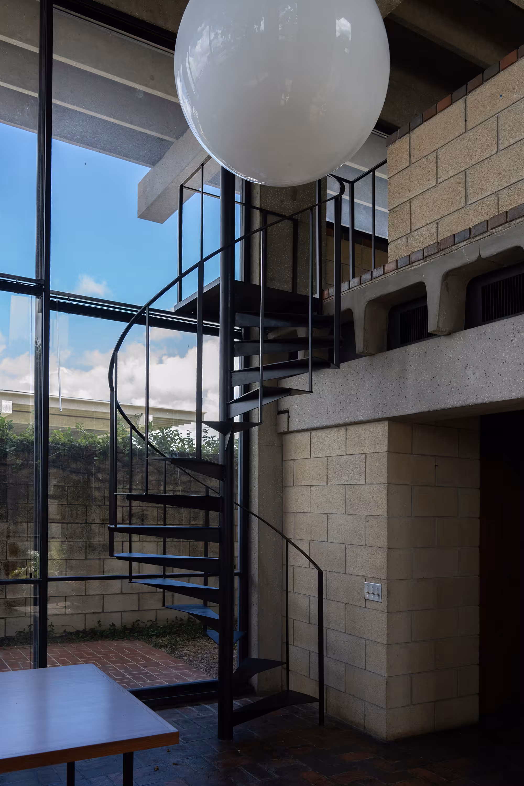 Interior view of a modernist space with exposed concrete beams and a dramatic black spiral staircase ascending beside a large globe light fixture, framed by tall glass walls that open to a private courtyard. {vertical}
