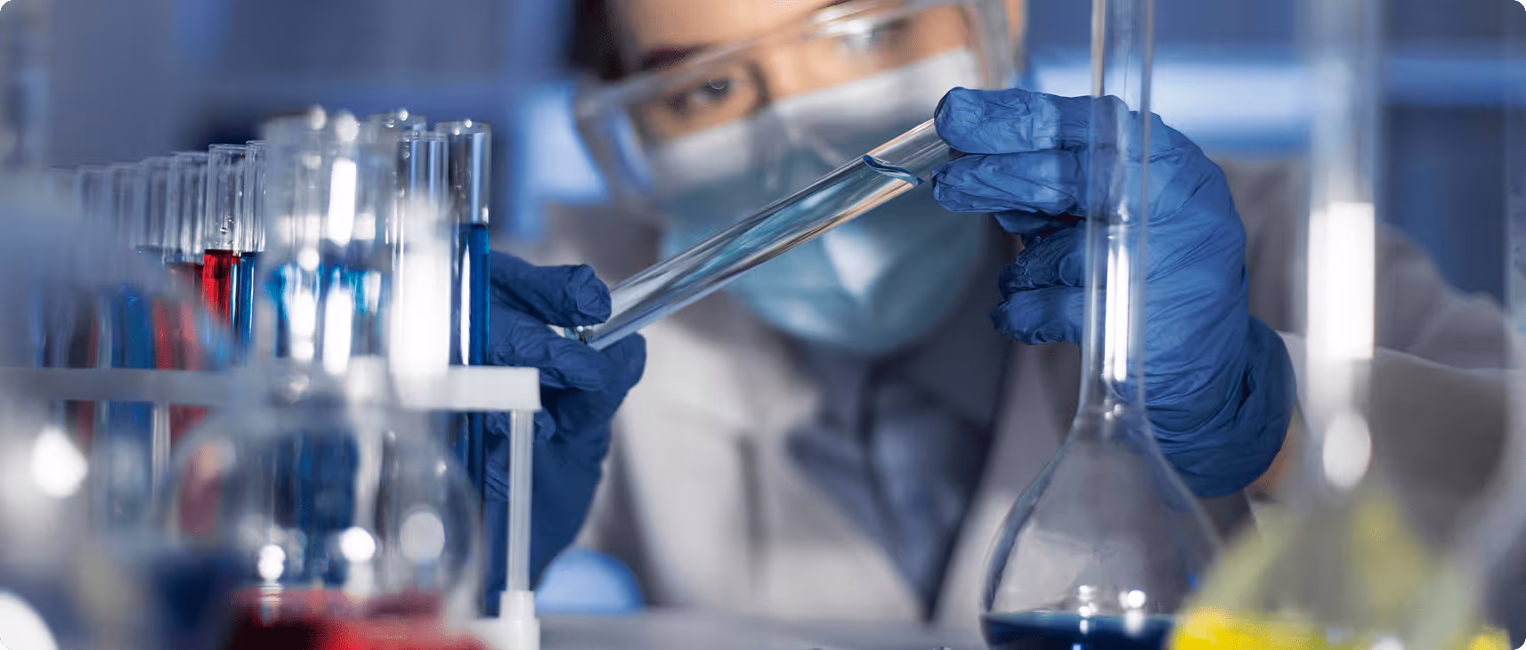Scientist wearing gloves, mask, and goggles examining a test tube in a laboratory, surrounded by various colored chemical samples—representing molecular research or new compound development.