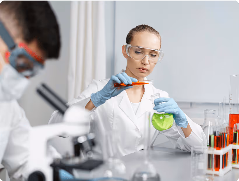 A female scientist in a lab coat and safety goggles conducts a chemical experiment, holding a flask of green liquid while using a pipette, with another scientist working in the background.