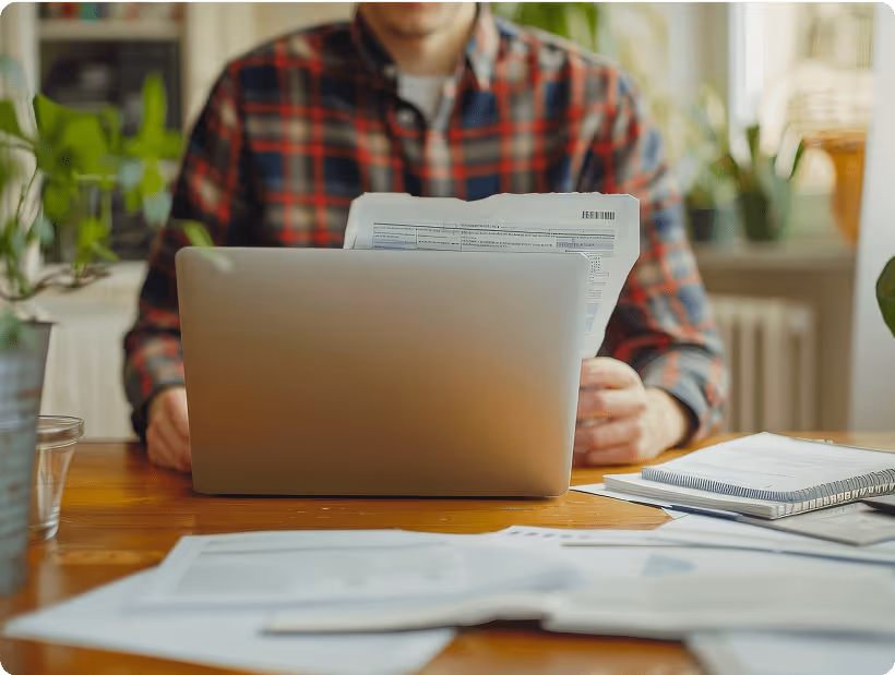 Man reviewing printed documents while working on a laptop at a wooden desk, surrounded by paperwork and plants.