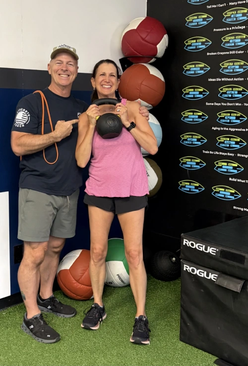 Fitness coach and client smiling while posing with a kettlebell in a gym.
