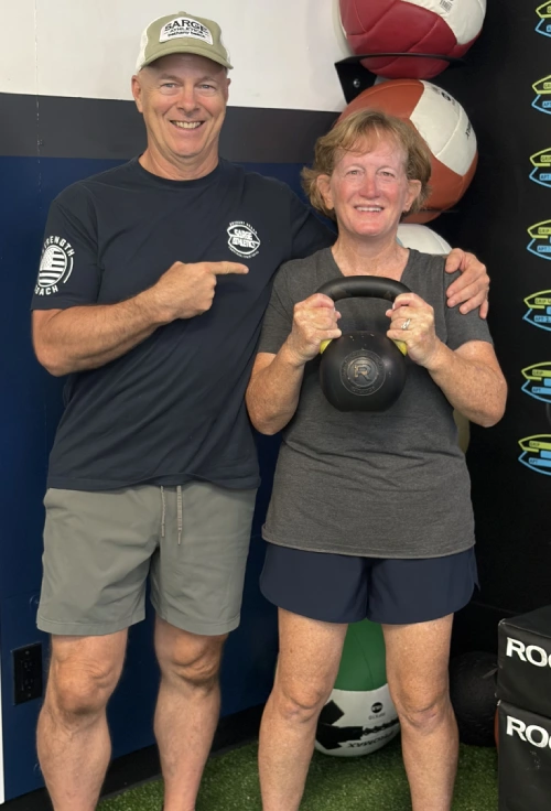 Fitness coach posing with a client holding a kettlebell in a gym.